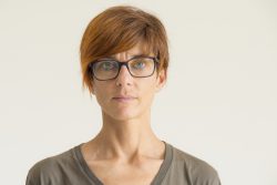 Waist up portrait of mature woman with red hairs, green eyes, eye glasses and serious facial expression, standing against the wall. Natural soft daylight, natural skin, neutral background.